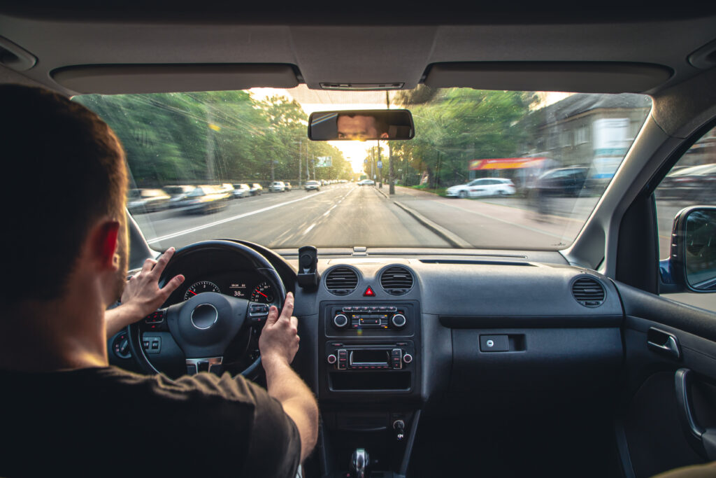 View from the back seat of a driver navigating a modern car on a city road during sunset, showing the dashboard and steering wheel.