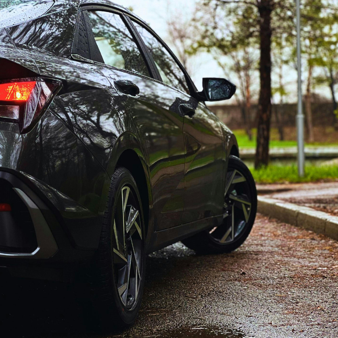 Rear three-quarter view of a dark grey modern sedan parked on a wet pavement, highlighting sharp body lines, a glowing red taillight, and two-tone alloy wheels.