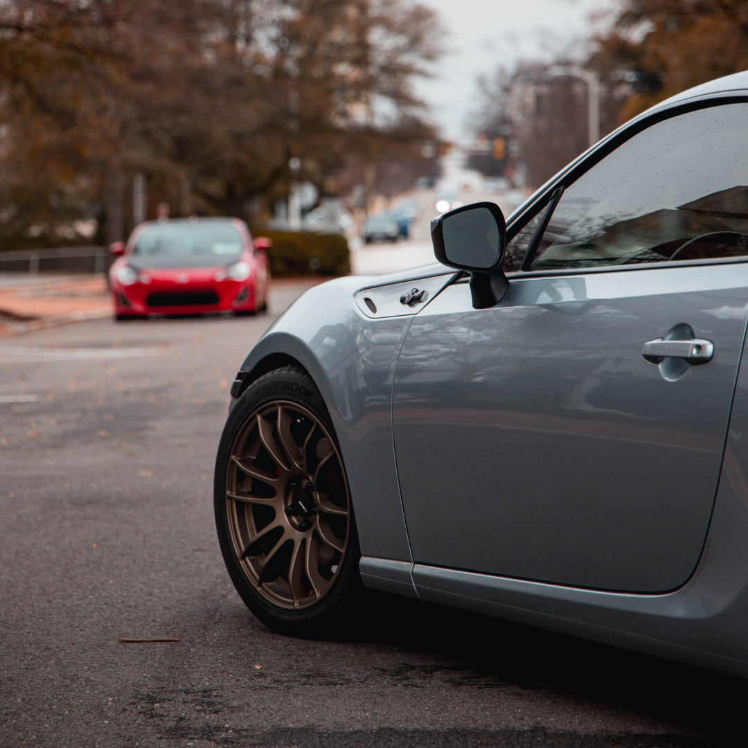 Close-up side profile of a silver-grey sports coupe with bronze multi-spoke aftermarket wheels, parked on a street with a red sports car in the blurred background.