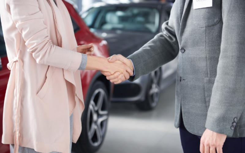 A professional handshake between a salesperson in a grey blazer and a client in a light-colored coat in front of a red vehicle at a car dealership.