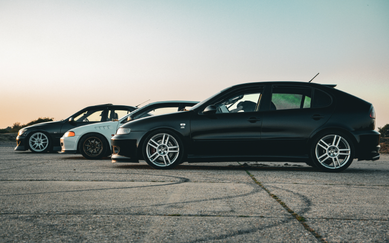 Profile view of three modified hatchbacks and sedans—one matte black, one white, and one glossy black—parked in a row on an open asphalt lot during sunset.