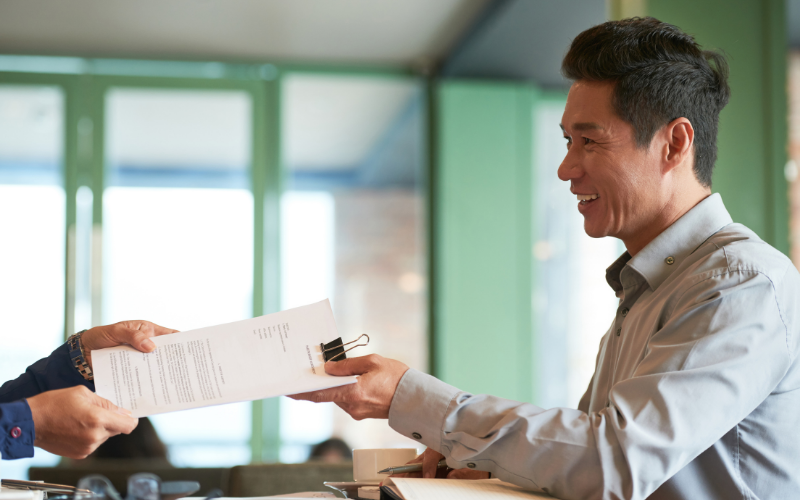 A smiling professional man in a grey collared shirt receiving a paper document or bid offer from another person in an office setting.