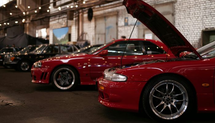 A side profile of multiple red sports cars with open hoods, showcasing high-performance engines and aftermarket wheels in a well-lit indoor warehouse.